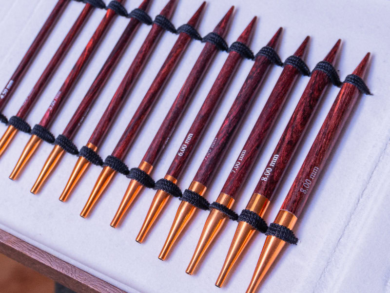 Un ensemble d'aiguilles à tricoter en bois à pointes en cuivre, disposées en rangée sur une surface blanche. Chaque aiguille, comme celles des meilleures aiguilles circulaires interchangeables, est marquée de sa taille et maintenue par des élastiques noirs.