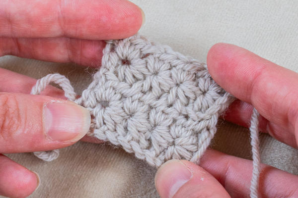 Gros plan de deux mains tenant un petit morceau carré de tissu crocheté avec du fil gris clair, présentant une texture détaillée et des points utilisant des fantaisies au crochet.