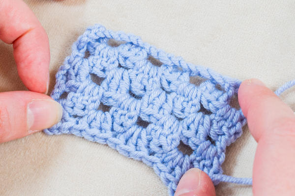 Deux mains tiennent un petit morceau de tissu bleu au crochet, présentant un motif de points ouverts - un exemple de fantaisies au crochet - sur une surface de couleur claire.