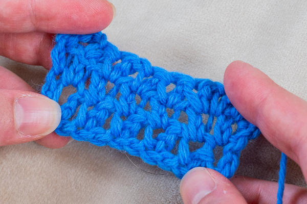 Deux mains tiennent un petit morceau de tissu bleu crocheté avec un motif ouvert et texturé, mettant en valeur des points fantaisies au crochet, sur un fond beige.
