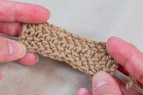 Deux mains tiennent un petit morceau de tissu crocheté marron clair, montrant la texture et les points fantaisies au crochet sur un fond neutre.