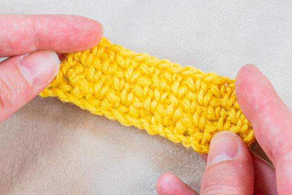Deux mains tiennent un petit morceau rectangulaire de tissu crocheté jaune vif sur un fond clair, mettant en valeur les points fantaisies au crochet. Les points sont clairement visibles.