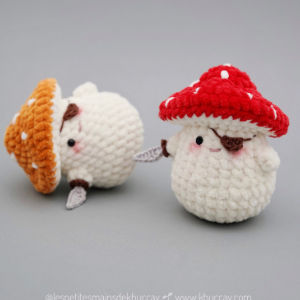 Deux adorables champignons amigurumi gratuits avec des visages souriants ; l'un a un chapeau orange et est couché sur le côté, l'autre se tient debout avec un chapeau rouge et des taches blanches, tous deux tenant de minuscules épées grises.
