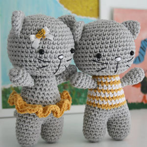 Deux poupées-chats gris crochetées en amigurumi gratuit se tiennent côte à côte. L'une porte une jupe jaune avec une petite abeille sur la tête, tandis que l'autre porte un haut rayé jaune et blanc. Des motifs colorés sont flous en arrière-plan.
