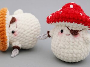 Deux créatures en peluche amigurumi crochet gratuit : l'une avec un chapeau de champignon rouge et une petite épée debout, et l'autre allongée avec un chapeau orange - toutes deux ont un corps blanc texturé et des joues rougissantes, reposant sur une surface grise.