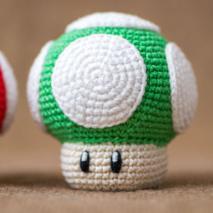 Une peluche amigurumi gratuite ressemblant à un champignon vert et blanc avec deux yeux noirs, inspiré du champignon 1-Up de la série de jeux vidéo Mario, repose sur une surface marron.