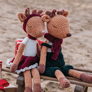 Deux poupées de rennes crochetées sont assises côte à côte sur un banc en bois. L'une porte une jupe et un bandeau rouges, l'autre un pantalon vert et une écharpe rouge. Toutes deux affichent une expression détendue. Une source d'inspiration idéale pour des projets d'animaux au crochet gratuits à télécharger.