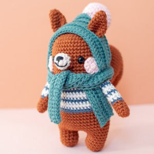 Un jouet écureuil brun au crochet portant un pull rayé bleu, une écharpe bleu sarcelle et une capuche bleu sarcelle avec un pompon blanc se dresse sur un fond pêche - une inspiration parfaite pour les animaux au crochet gratuits à télécharger.