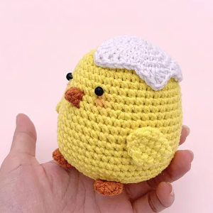 Une main tient un petit poussin jaune crocheté avec une coquille d'œuf blanche sur la tête, un bec et des pattes orange et des yeux en perles noires sur un fond rose clair - parfait pour ceux qui recherchent des animaux au crochet gratuits à télécharger.