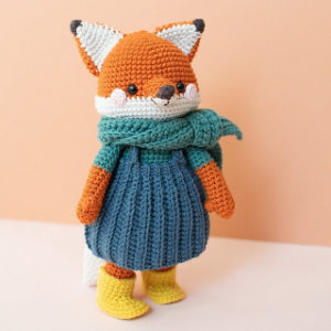 Une poupée renard crochetée se tient debout, vêtue d'une robe bleue, d'une écharpe verte et de bottes jaunes, sur un fond couleur pêche ; une inspiration parfaite pour des animaux au crochet gratuits à télécharger. Le renard a une fourrure orange, des joues blanches et des oreilles à bouts noirs.