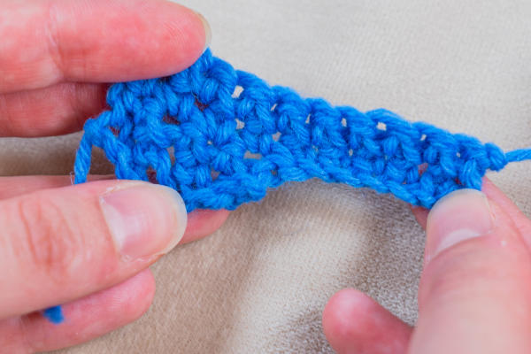 Gros plan des mains tenant un petit morceau de tissu bleu au crochet, mettant en évidence les points individuels et la texture avec des augmentations visibles au crochet sur un fond beige clair.