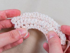Gros plan de deux mains tenant un petit morceau incurvé de tissu crocheté de couleur claire sur un fond beige, mettant en évidence de délicates augmentations au crochet.