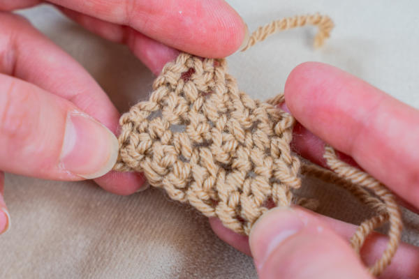 Deux mains tenant un petit morceau d'ouvrage au crochet en fil fauve, mettant en évidence les augmentations au crochet et montrant les points et la texture de près sur un fond neutre.
