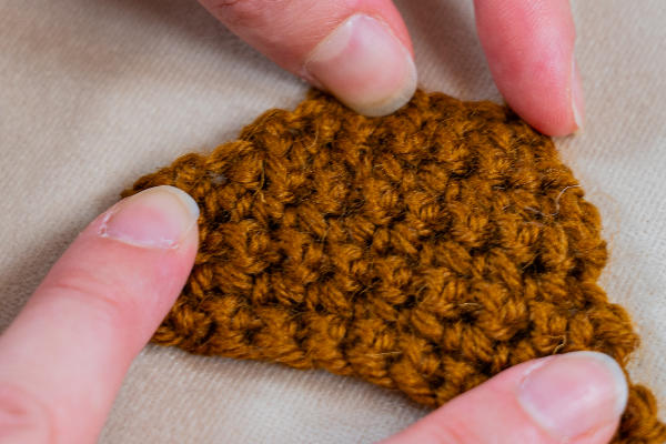 Gros plan de deux mains tenant un morceau triangulaire de tissu crocheté marron sur une surface beige, mettant en valeur de délicates augmentations au crochet.