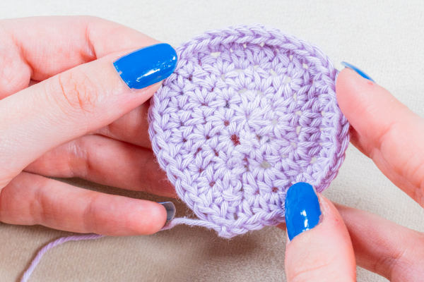 Une personne avec du vernis à ongles bleu vif tient un morceau rond et plat d'un ouvrage au crochet violet clair, montrant les points détaillés et les augmentations nettes pour crocheter en rond.