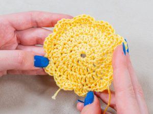 Deux mains au vernis à ongles bleu tiennent une fleur jaune crochetée, dont le bord festonné présente des augmentations pour crocheter en rond, sur un fond neutre.