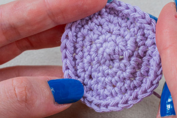 Gros plan sur des mains tenant un morceau rond de crochet violet clair, avec les ongles bleu vif de la personne visible - montrant les augmentations pour crocheter en rond pour des motifs circulaires magnifiquement détaillés.