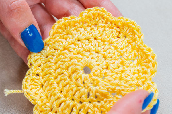 Une personne aux ongles peints en bleu tient un morceau de fil jaune crocheté en forme de fleur aux bords festonnés, présentant des augmentations pour crocheter en rond afin d'ajouter de la texture et de la dimension.