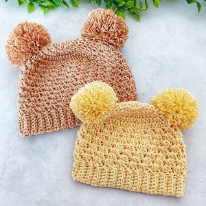 Deux bonnets pour bébé au crochet, l'un marron clair et l'autre jaune, chacun avec deux gros pompons sur le dessus, sont exposés sur une surface claire avec des feuilles vertes en arrière-plan, parfaits comme un charmant bonnet pour bébé au crochet.