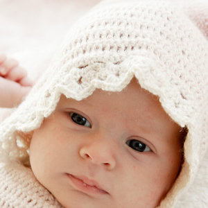 Gros plan d'un bébé à la peau claire portant un bonnet pour bébé au crochet de couleur crème, regardant doucement la caméra avec une expression calme.
