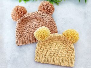Deux bonnets pour bébé au crochet avec des pompons sur le dessus, l'un en marron clair et l'autre en jaune, sont placés côte à côte sur une surface de couleur claire avec des feuilles vertes visibles en arrière-plan.