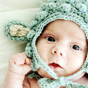 Un bébé portant un bonnet pour bébé au crochet avec des détails en forme d'oreilles regarde vers le haut avec de grands yeux tout en étant allongé sur une couverture de couleur claire.