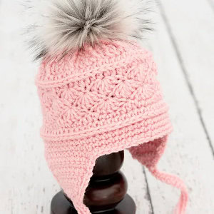 Un bonnet rose pour bébé au crochet avec des cache-oreilles et un pompon gris et blanc duveteux sur le dessus, présenté sur un support en bois foncé sur un fond en bois blanc.