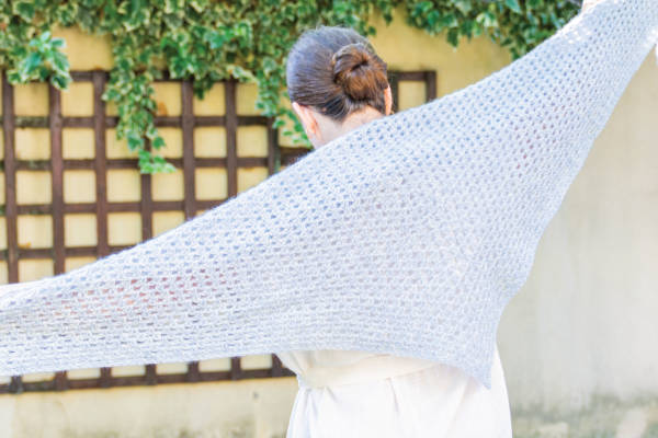 Une personne aux cheveux bruns coiffés en chignon se tient debout, dos à l'appareil photo, et tend un grand châle au crochet en forme de triangle gris clair. Du lierre vert grimpe sur un treillis et un mur à l'arrière-plan.