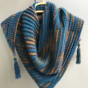 Un châle triangulaire tricoté en fil panaché bleu, gris et orange est suspendu à un cintre. Rappelant les châles au crochet, il présente des motifs texturés et des pompons ornés de perles à deux coins.