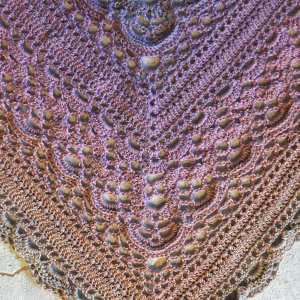 Gros plan d'un châle triangulaire, finement crocheté, aux couleurs dégradées de violet, rose et marron, avec des motifs texturés en relief et des détails en dentelle - un élégant exemple de l'artisanat des châles au crochet.