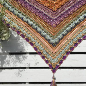 Gros plan de châles au crochet vibrants aux bords festonnés dans des tons orange, violet, vert et gris, drapés sur des lattes de bois blanches et terminés par un pompon à l'extrémité pointue.