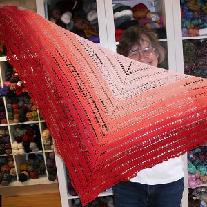 Une personne se tient devant des étagères remplies de fils colorés, tenant un grand châle au crochet triangulaire présentant un motif dégradé qui passe du rouge au rose clair.