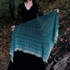 Une femme vêtue d'une longue robe en dentelle noire se tient debout dans un bois, tenant un grand châle bleu sarcelle en dentelle. Ce châle au crochet, aux motifs délicats et aux magnifiques dégradés de couleurs, s'ouvre gracieusement devant elle.
