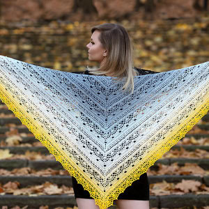 Une femme se tient dehors, sur des marches de pierre recouvertes de feuilles d'automne, tenant un grand châle triangulaire au crochet aux tons dégradés de bleu, blanc et jaune. Le châle en dentelle est largement étalé derrière elle.
