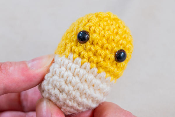 Une main tient un petit personnage jaune et blanc crocheté avec deux yeux noirs en plastique - illustrant la technique du changement de couleur au crochet - sur un fond uni.