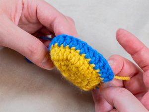 Deux mains tiennent un petit objet rond crocheté avec des points jaunes au centre et une bordure bleue - un exemple de la façon de changer de couleur au crochet - sur un fond neutre.