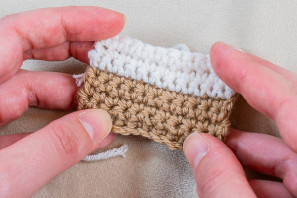 Deux mains tiennent un petit rectangle crocheté montrant comment changer de couleur au crochet, avec une partie inférieure beige et une partie supérieure blanche, sur un fond de tissu neutre.