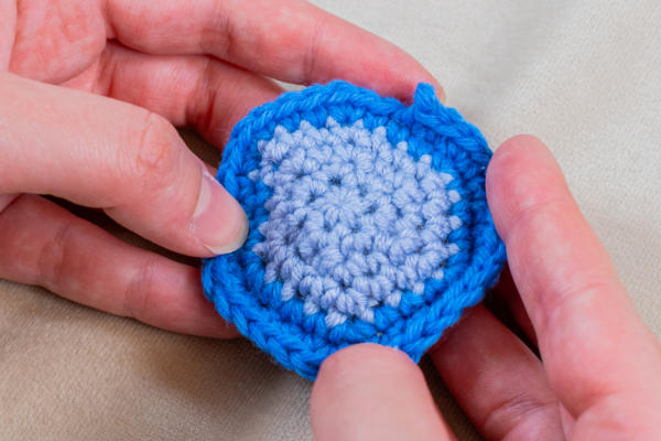 Gros plan des mains tenant une petite pièce ronde au crochet, montrant comment changer de couleur au crochet avec un centre bleu clair et une bordure bleu vif, sur un fond de tissu clair.