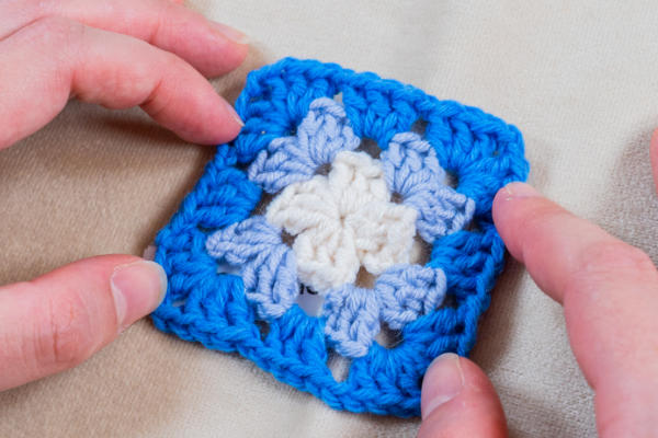 Deux mains tiennent un carré de grand-mère crocheté avec un centre blanc, un milieu bleu clair et une bordure bleu vif - parfait pour s'entraîner à changer de couleur au crochet - reposant sur un fond de tissu beige.