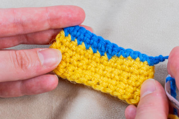 Gros plan sur des mains tenant une petite pièce crochetée à deux rangs : jaune vif en bas et bleu en haut, illustrant comment changer de couleur au crochet, sur un fond de tissu neutre.