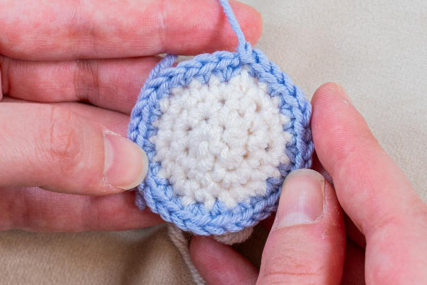Gros plan des mains tenant une petite pièce ronde au crochet avec un centre blanc et une bordure bleue, montrant la technique pour changer de couleur au crochet lorsque le fil bleu est ajouté pour continuer le projet.