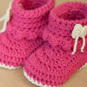 Une paire de chaussons bébé roses au crochet avec semelles blanches et petits noeuds blancs sur les côtés, posés sur une surface marron clair.