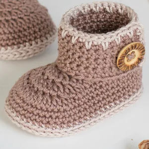 Un chausson bébé au crochet en fil marron clair et crème avec un bouton décoratif en bois sur le côté, placé sur une surface blanche - parfait comme adorable chaussons bébé au crochet pour votre tout-petit.
