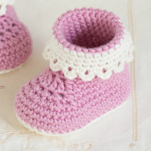 Chausson bébé rose au crochet, orné d'un bord festonné blanc décoratif, posé sur un fond en tissu clair. Un autre chausson bébé similaire au crochet est partiellement visible en arrière-plan.