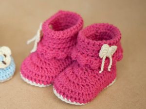 Adorables chaussons bébé roses au crochet avec semelles blanches et charmants nœuds blancs, joliment présentés sur une surface marron clair.