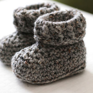 Une paire de chaussons bébé gris crochetés à la main est présentée sur une surface blanche. Ces chaussons bébé au crochet présentent un motif texturé et des revers repliés, ajoutant une touche de charme artisanal.