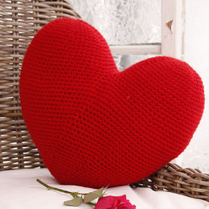 Un coussin rouge en forme de cœur, inspiré des cœurs en crochet, repose sur une chaise en osier, avec une rose rouge devant. Une fenêtre givrée complète ce décor chaleureux.