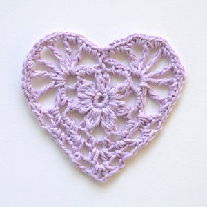 Un cœur délicat en crochet violet clair avec un motif floral complexe, présenté sur un fond blanc uni.