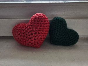 Deux cœurs en crochet, l’un rouge et l’autre vert foncé, reposent côte à côte sur une surface en bois près d’une fenêtre baignée de douce lumière naturelle.