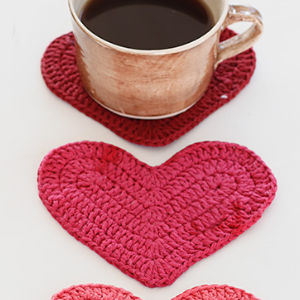 Une tasse en céramique marron remplie de café repose sur un sous-verre en forme de cœur crocheté rouge, avec deux autres cœurs en crochet rose et rouge placés à proximité sur une surface blanche.